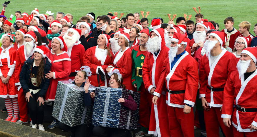 Crowd of people dressed as Santa smiling, two people wrapped as presents at the front of the crowd