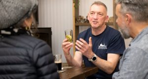 Male head brewer, glass in hand looking forwards, sat at a table talking to two people facing him at the brewery bar