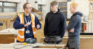 Joinery lecturer in orange jacket teaching two white male students, wooden table and tools in front