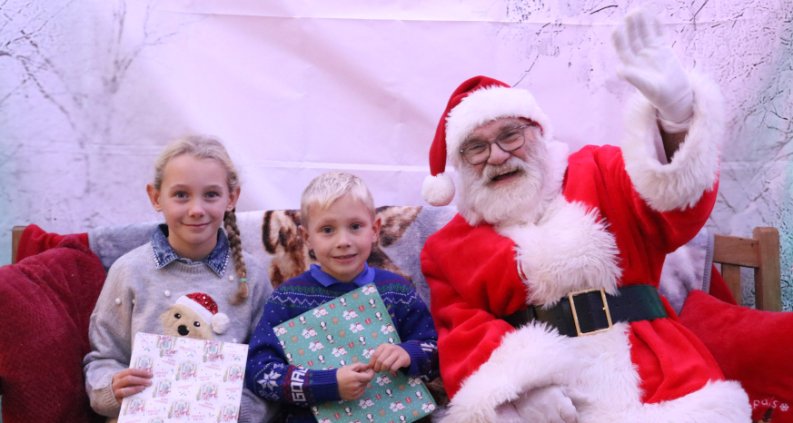 Two young children with wrapped gifts sat on the left of Santa, waving