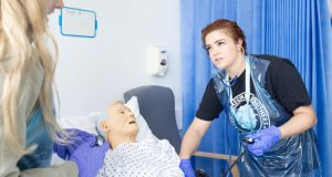 White student nurse practicing using a stethoscope on a dummy in hospital bed