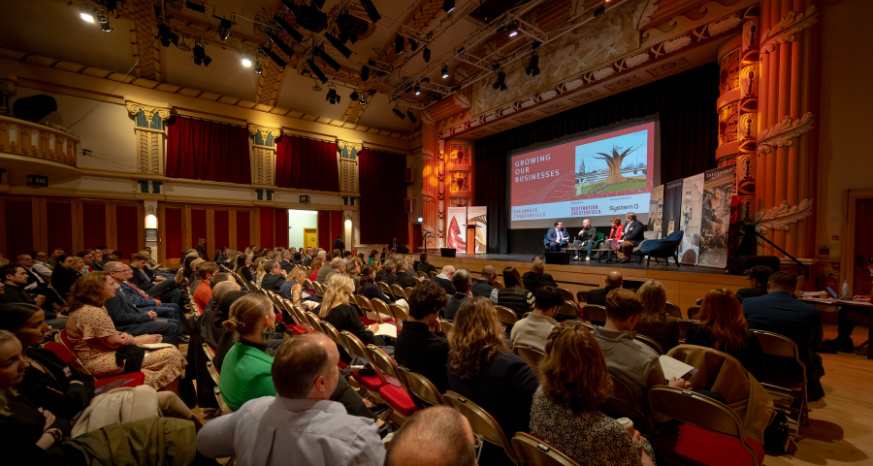 Large audience looking at slide that says 'growing our businesss' at the celebrate chesterfield 2024 conference whilst panel with Adam Doyle, Josh Marsh, Mike Isherwood and Cllr Tricia Gilby