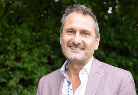 Man with short hair and pastel pink jacket stood in front of dark green foliage background