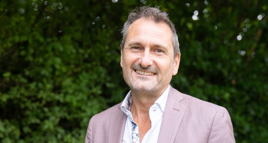 Man with short hair and pastel pink jacket stood in front of dark green foliage background