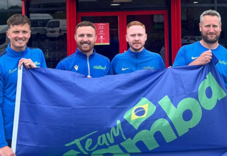 4 males holding a giant flag that reads #team samba - the 4 owners of samba sports coaching