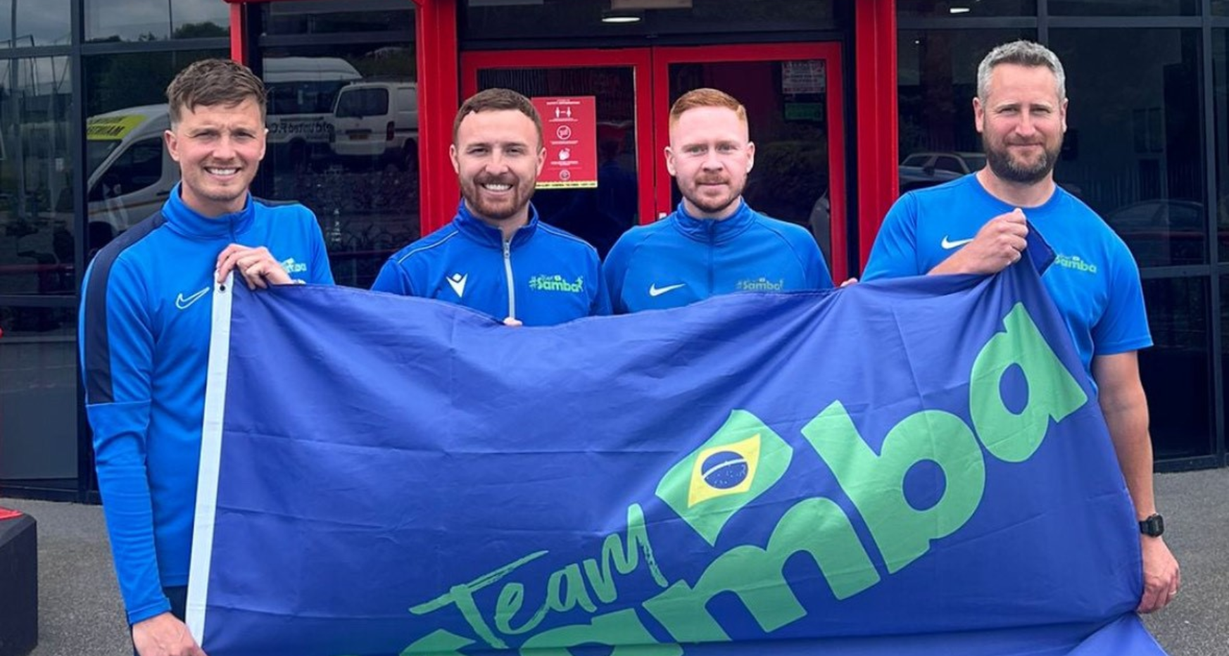 4 males holding a giant flag that reads #team samba - the 4 owners of samba sports coaching