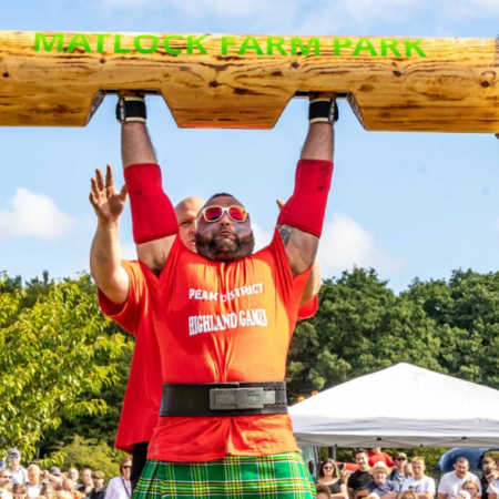 A man lifts a huge log above his head while a crowd watches on.